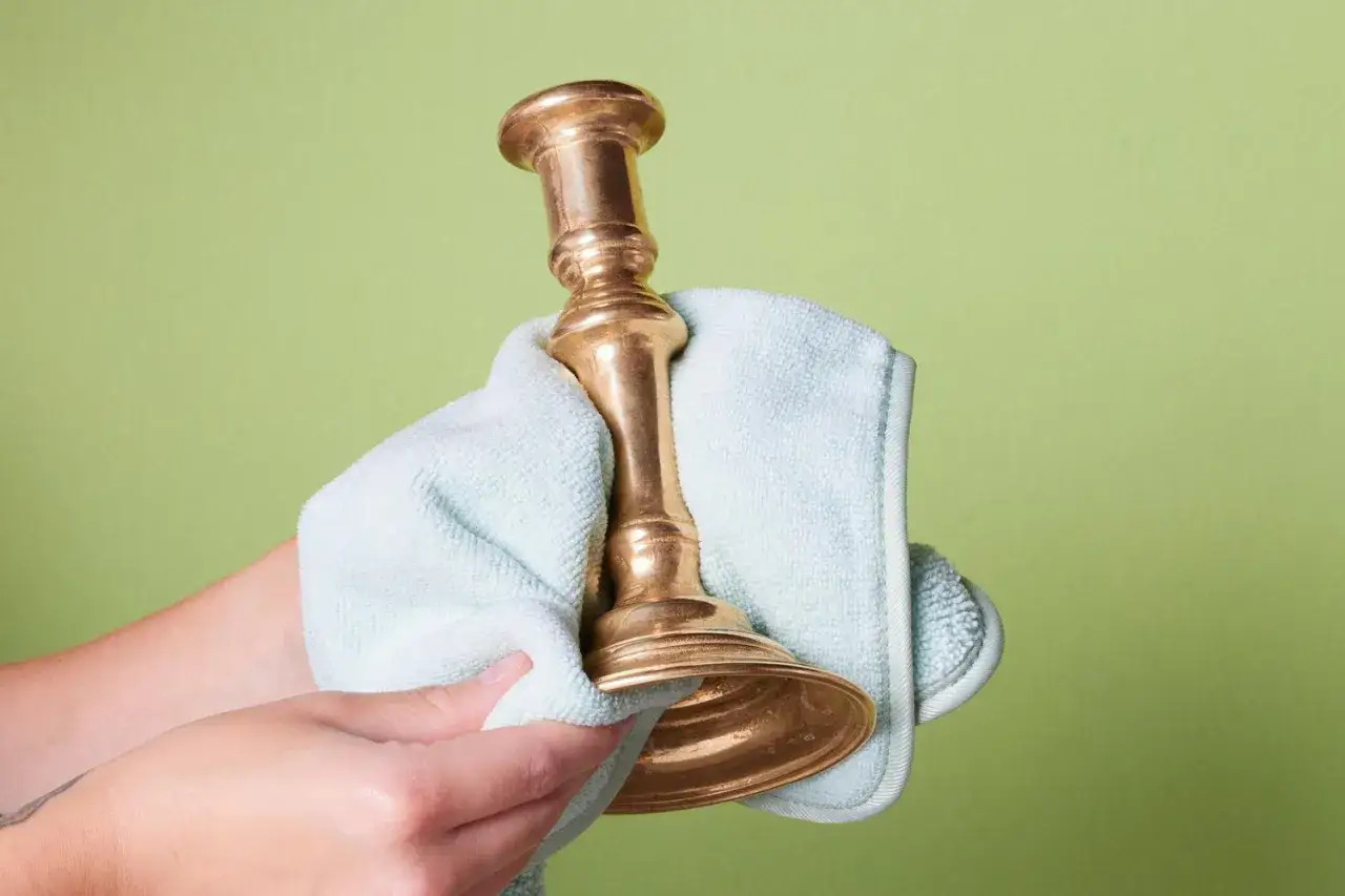 A hand uses a light blue cloth to polish a vintage brass candlestick, showing how to polish brass to a shine.