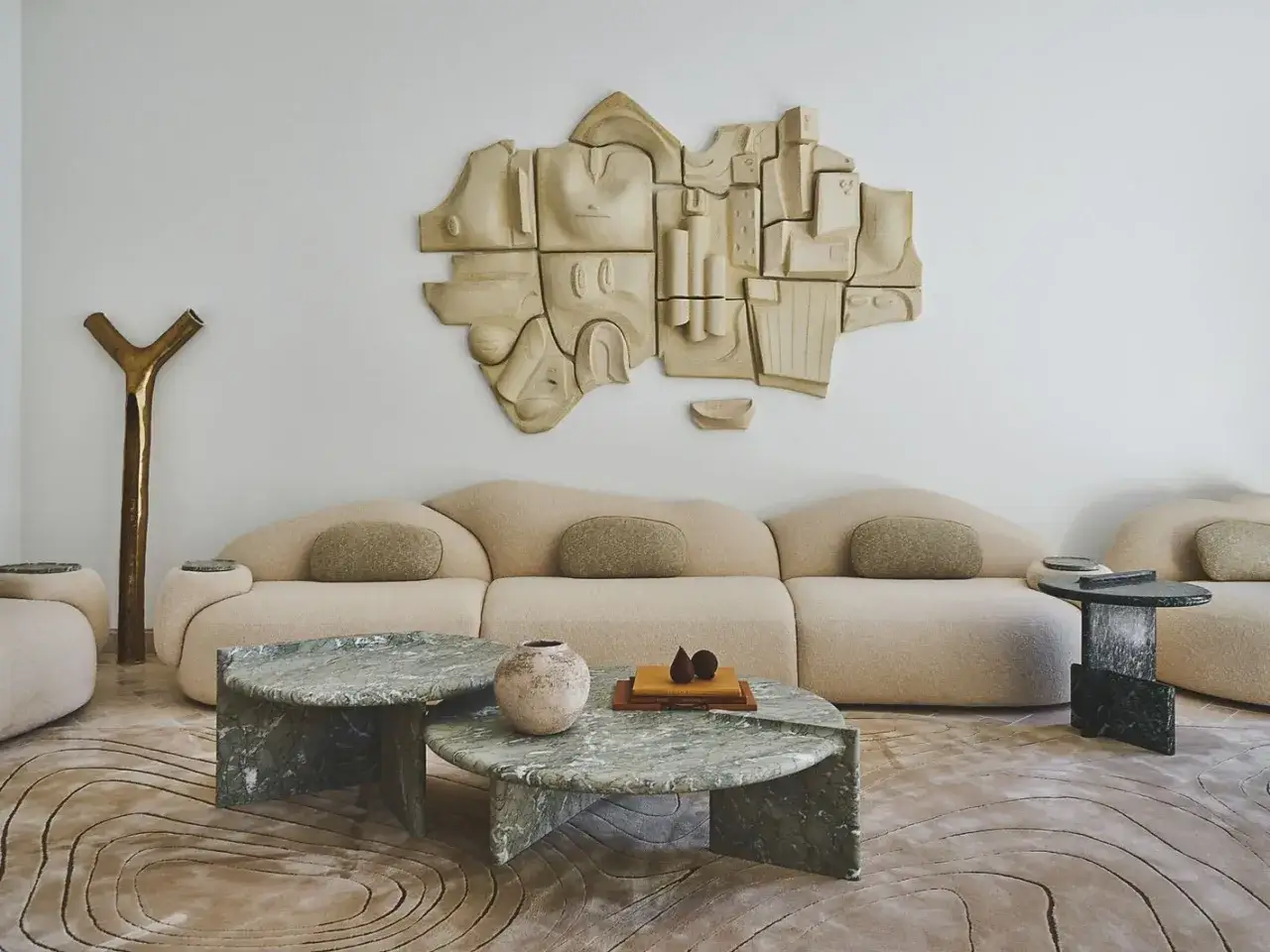 Une sculpture abstraite en terre cuite vient habiller un mur blanc, dominant un canap&eacute; beige aux formes douces et des tables basses en marbre vert.