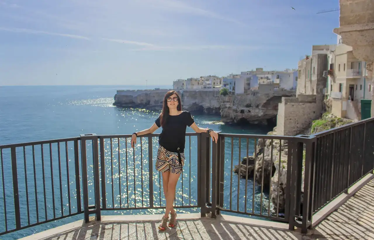 Donna sorridente con vista mozzafiato sul mare a Polignano a Mare, Italia. Le case bianche aggrappate alla scogliera creano uno scenario da cartolina.