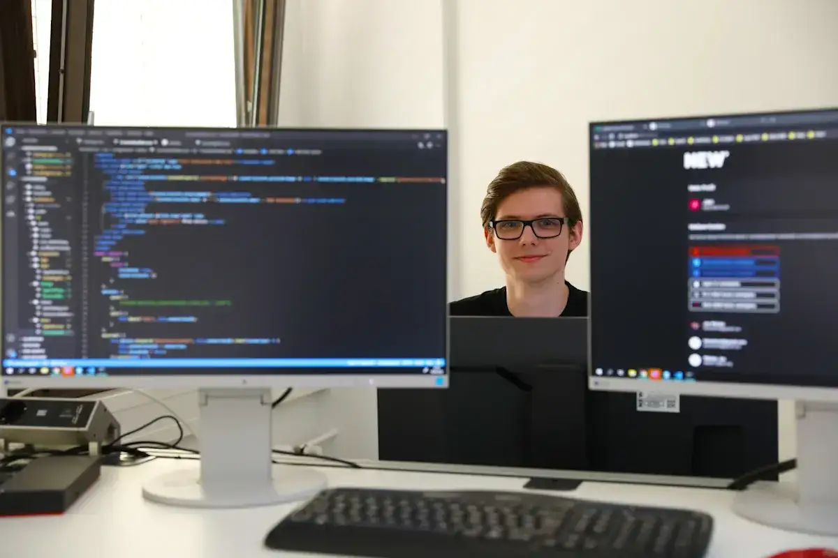 Ein junger Mann mit Brille l&auml;chelt in die Kamera, w&auml;hrend er als Werkstudent IT an zwei Bildschirmen mit Code arbeitet.