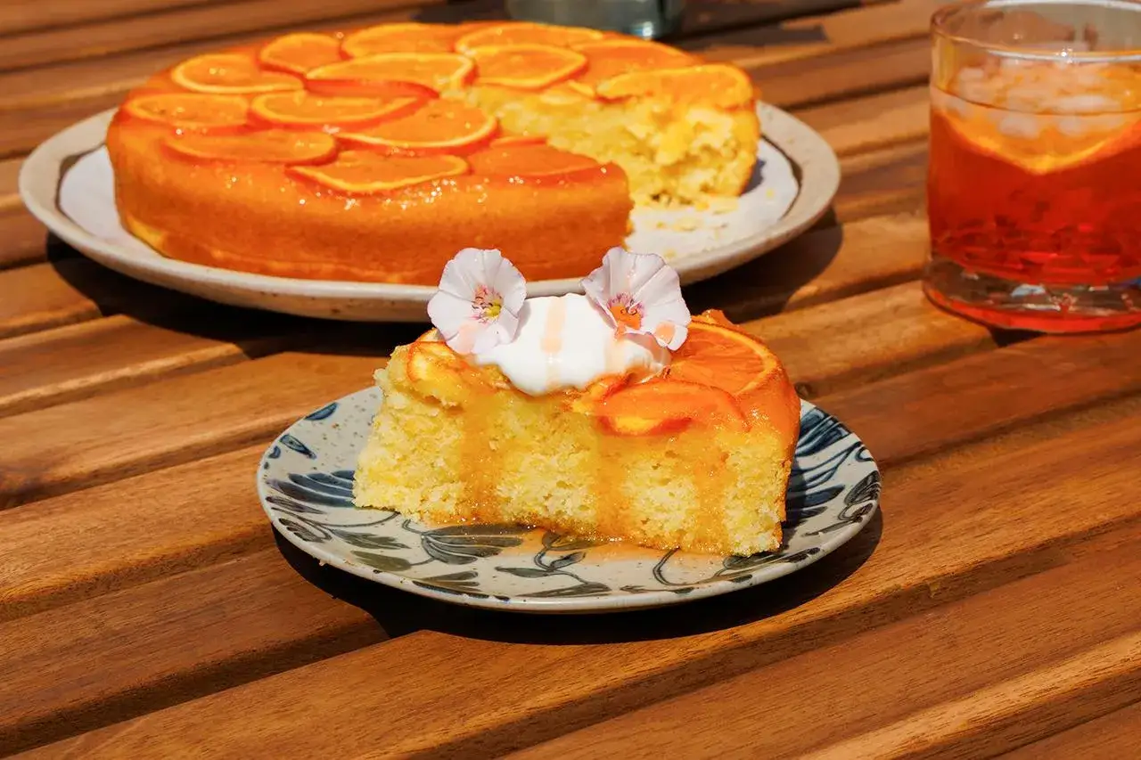 Saftige Aperol Torte mit Orangen, garniert mit Sahne und Blüten. Ein Stück auf einem Teller, daneben ein Glas Aperol Spritz.