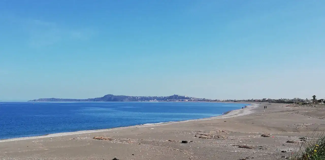Spiaggia di **Barcellona Pozzo di Gotto** con il mare blu intenso e la costa in lontananza sotto un cielo limpido.