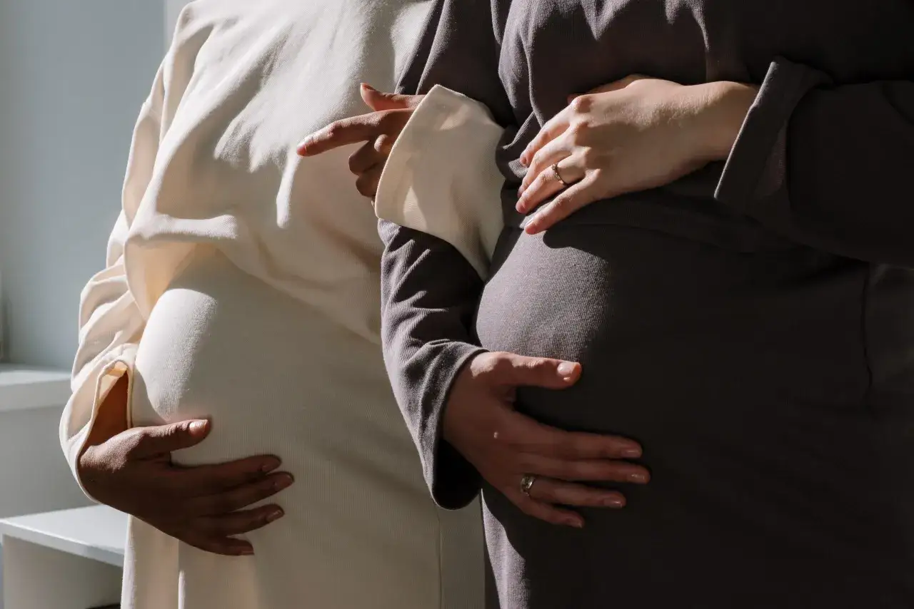 Due donne incinte si abbracciano, una con un vestito chiaro, l'altra con uno scuro. Si tengono la pancia, un gesto che ricorda che piangere in gravidanza fa male.