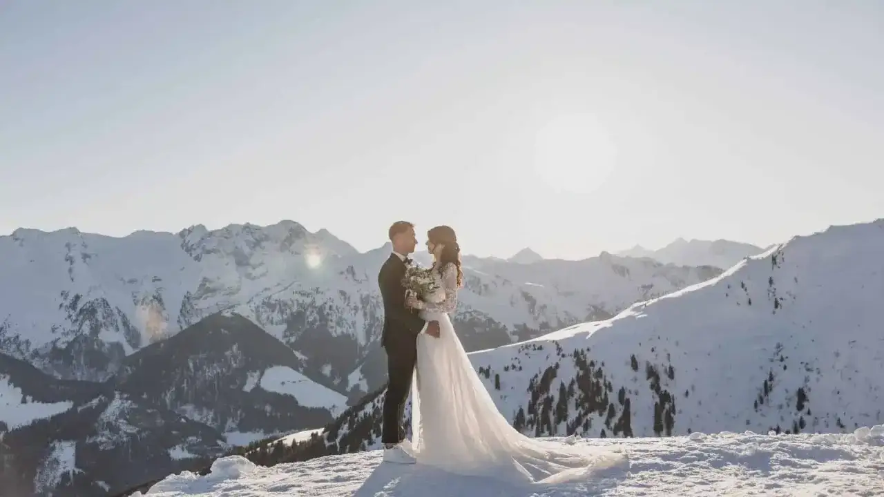 Ein Brautpaar steht sich in der verschneiten Berglandschaft gegenüber. Die Sonne scheint hinter ihnen, und die Berge sind mit Schnee bedeckt. Eine romantische Hochzeit im Schnee.