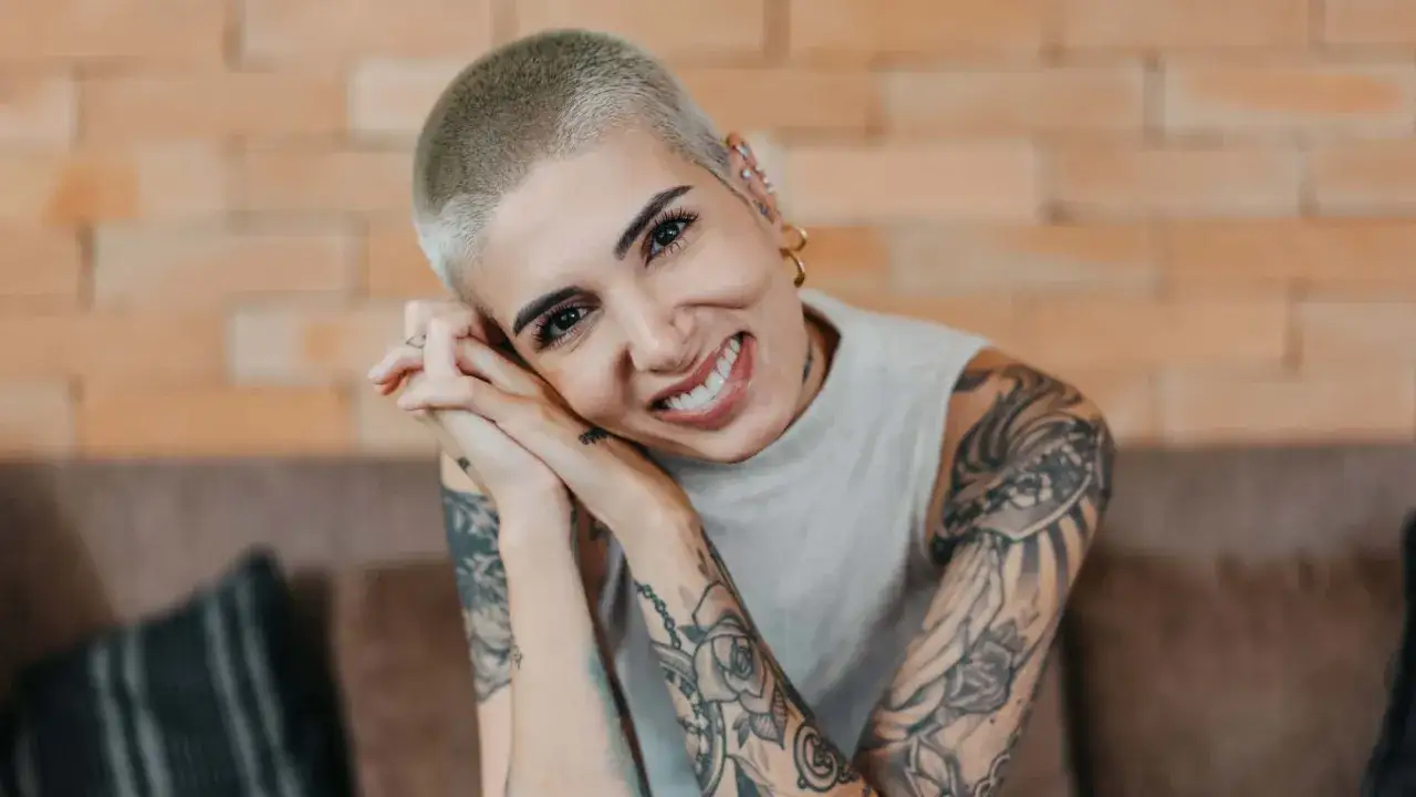 Lächelnde Frau mit Buzz Cut und vielen Tattoos, die ihre Hände vor dem Gesicht verschränkt.