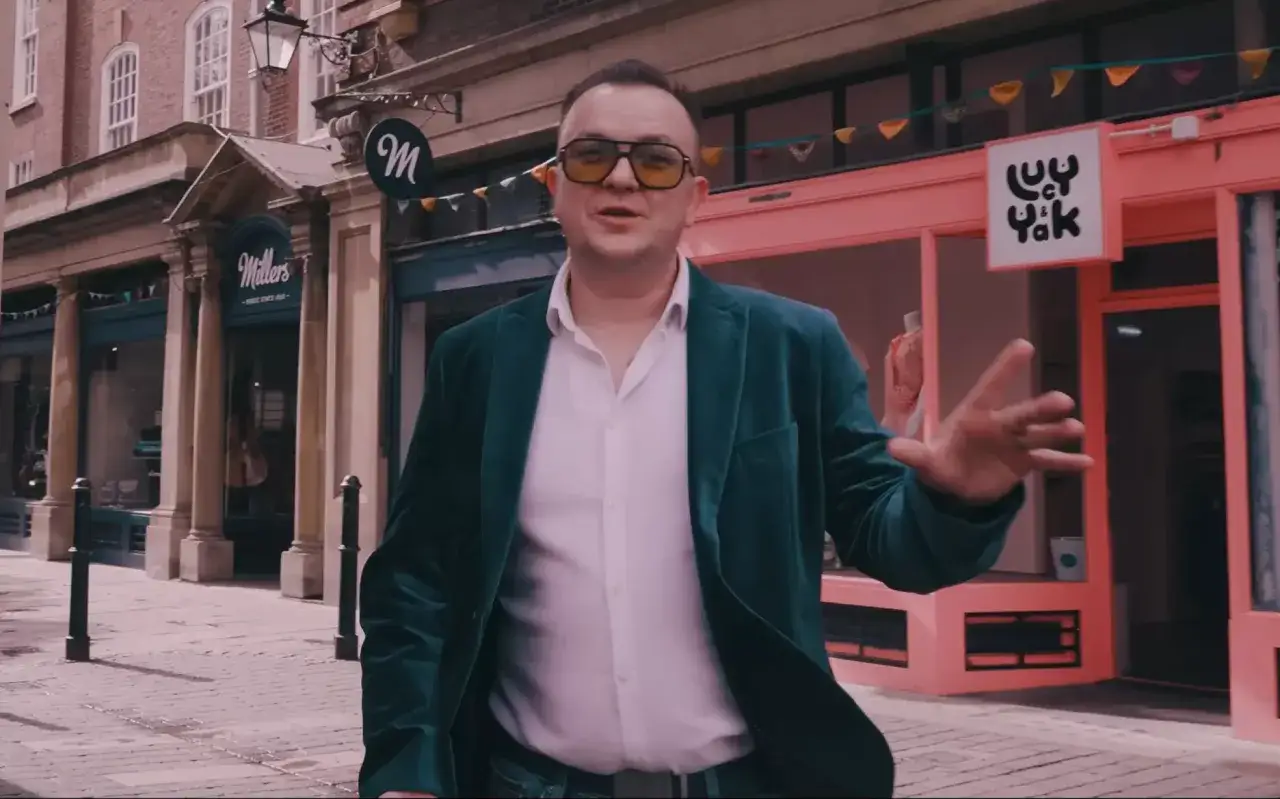 Mężczyzna w zielonym żakiecie i okularach opowiada o **pulsie miasta tekst**, stojąc przed sklepami.