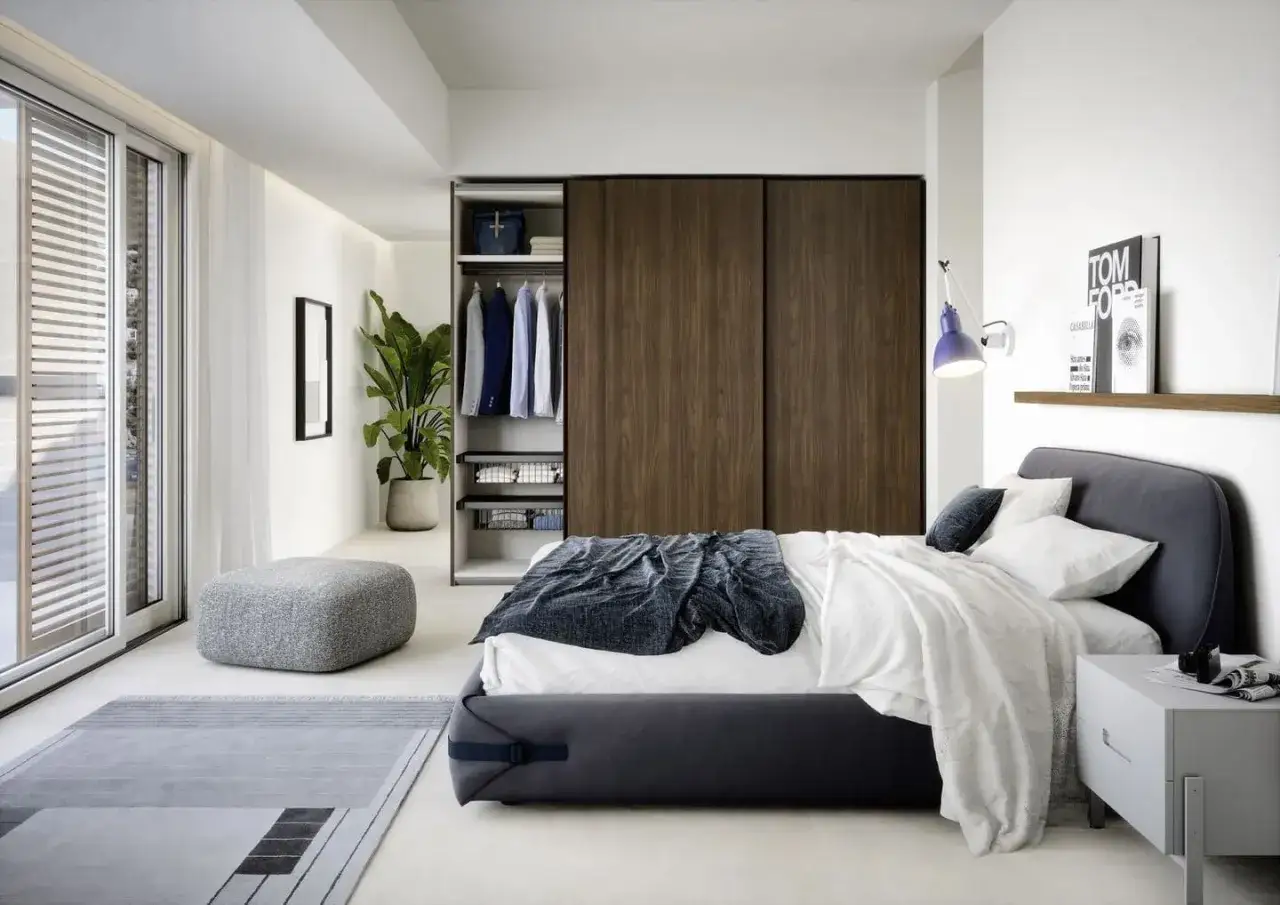 Ecco come arredare camera da letto: letto moderno, armadio a muro in legno, pouf grigio e pianta verde.