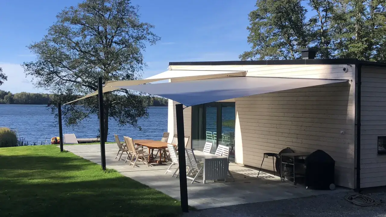 Solsegel ger skugga över uteplatsen vid sjön. Trädgårdsmöbler och grill står redo för sommarens middagar.