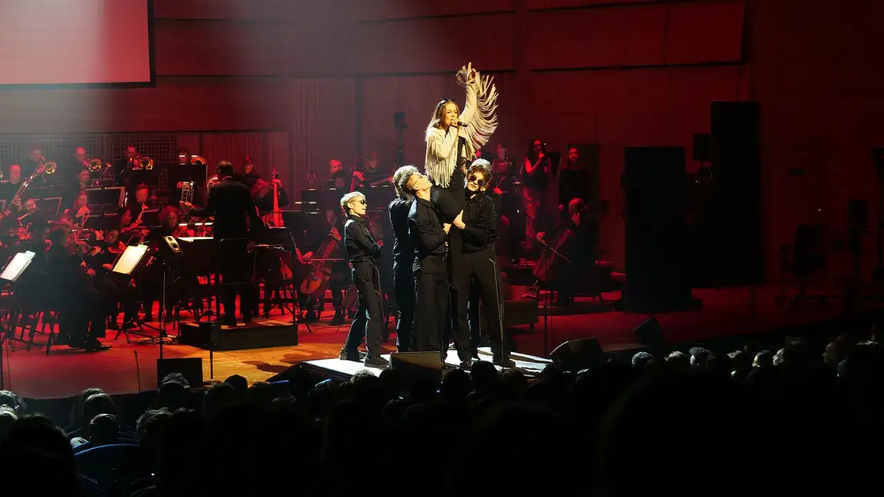 En ung artist sjunger på scenen under ett de geerhallen evenemang, omgiven av en orkester och publik.