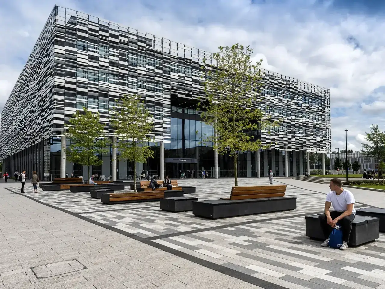 Façade moderne de l'université métropolitaine de Manchester. Des étudiants profitent du soleil sur des bancs extérieurs.