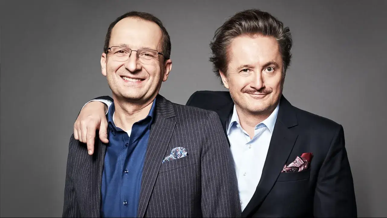Dwóch mężczyzn w garniturach, uśmiechniętych, z ręką na ramieniu. To piękni i bezrobotni obsada, gotowi na nowe wyzwania.