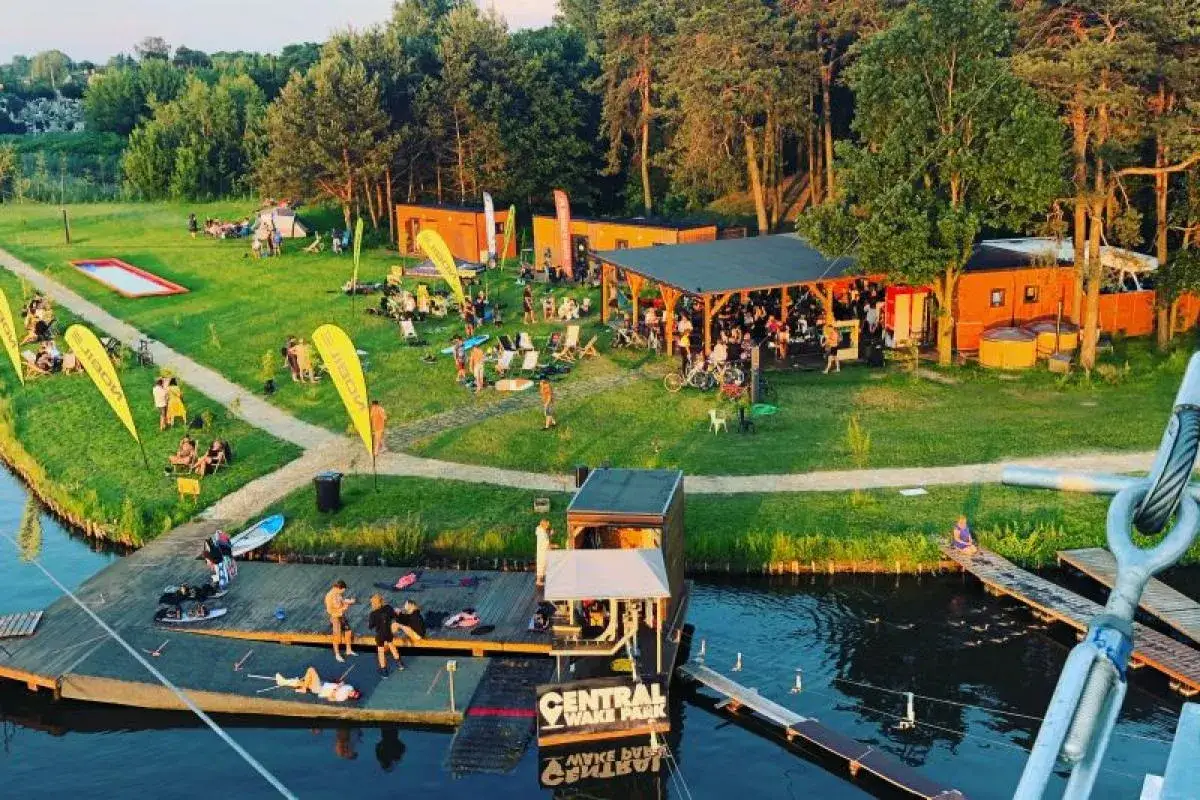 Letni dzień nad zalewem Mrożyczka. Ludzie odpoczywają na trawie, przy barze i na pomoście. W tle las i budynki Central Wake Park.