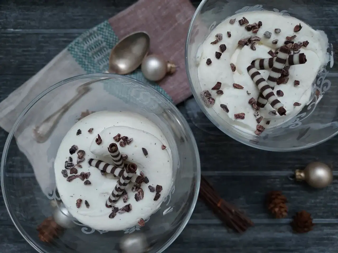 Deux coupes de chantilly chocolat blanc, parsemées de cacao et décorées de rouleaux de chocolat. Une ambiance festive avec des boules de Noël et des pommes de pin.