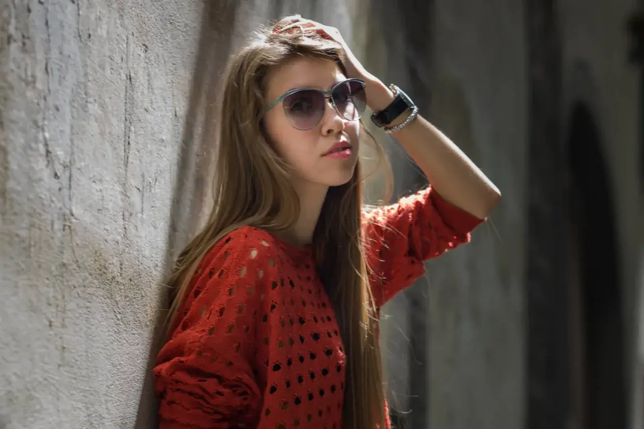 A young woman with long blonde hair, wearing sunglasses and a red sweater, leans against a textured wall. The portrait, captured with a Leica 75mm Summicron, highlights her features.