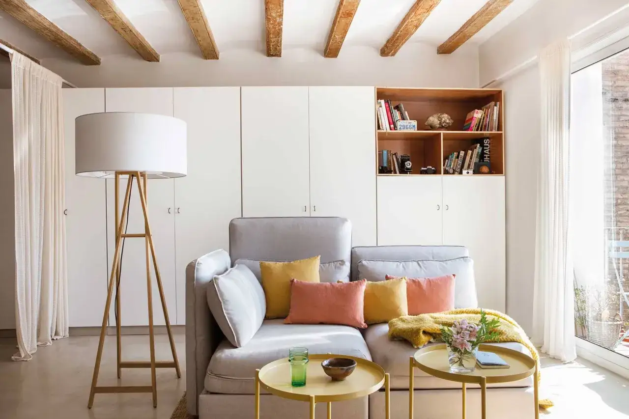 Ideas para decorar piso alquiler low cost: sofá gris, cojines coloridos, mesa auxiliar dorada, lámpara de pie y estantería con libros.