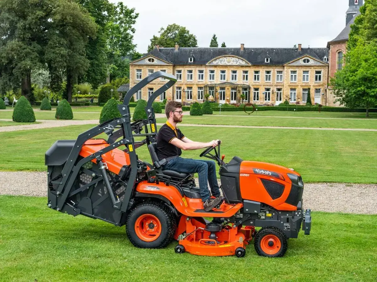 Mężczyzna na trawniku prowadzi pomarańczowy traktorek kosiarkę diesel Kubota G261, przygotowując się do koszenia przed okazałym budynkiem.