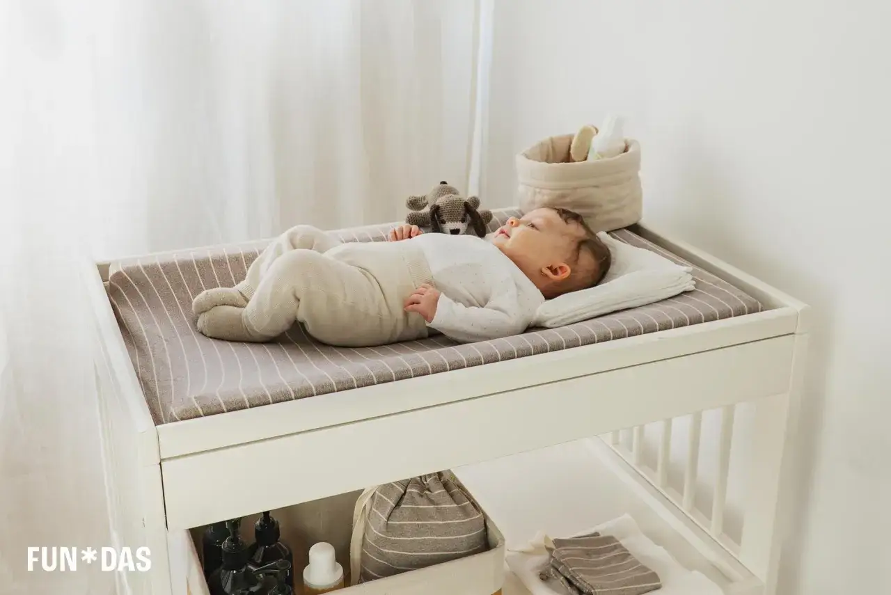 Beb&eacute; despierto en cambiador, parece que mi bebe tarda mucho en dormirse. Juguete de perro y cesta con productos.