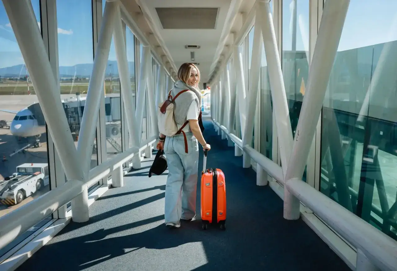 Frau mit orangefarbenem Koffer und Hut geht durch einen Flugsteig. Hoffentlich verpasst sie nicht den Anschlussflug!