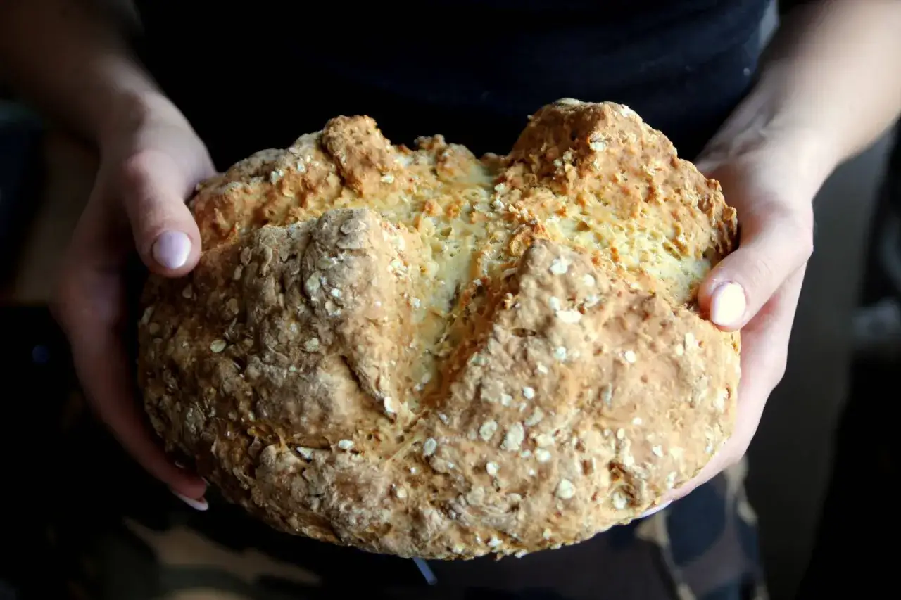 Ręce trzymają okrągły, złocisty bochenek soda bread, posypany płatkami owsianymi.