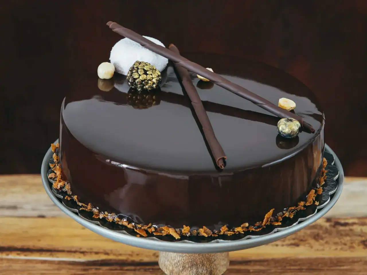 Torta cioccolatosa goduriosa con glassa lucida, decorata con riccioli di cioccolato, nocciole e una sfera dorata.