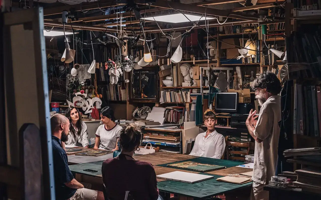 Un gruppo di studenti ascolta un artista nel suo atelier a Venezia. L'ambiente è ricco di opere e materiali artistici.