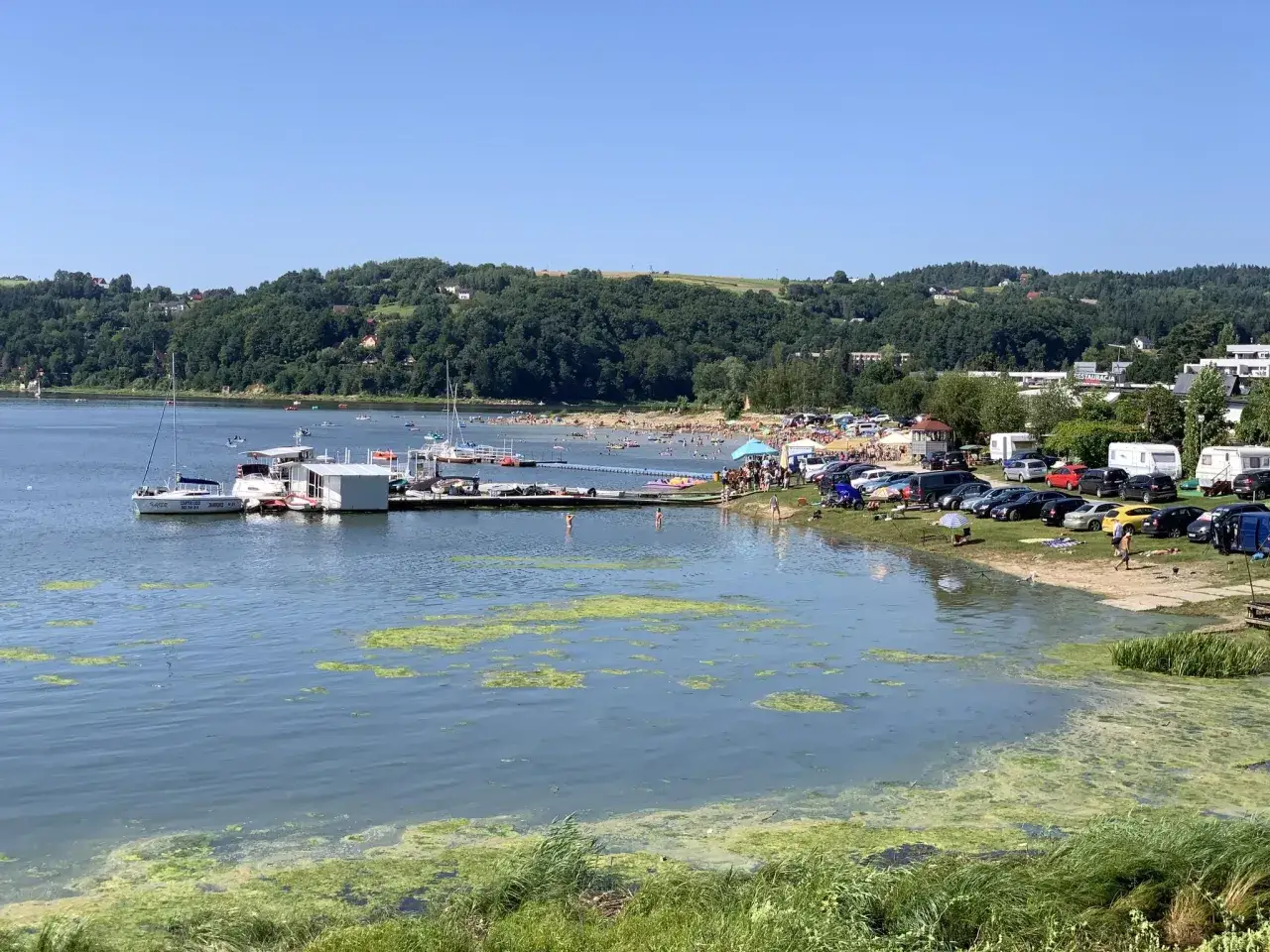 Jezioro Rożnowskie tętni życiem. Widać plażowiczów, łodzie, kampery i parking pełen samochodów. To idealne miejsce na letni wypoczynek i korzystanie z licznych atrakcji.