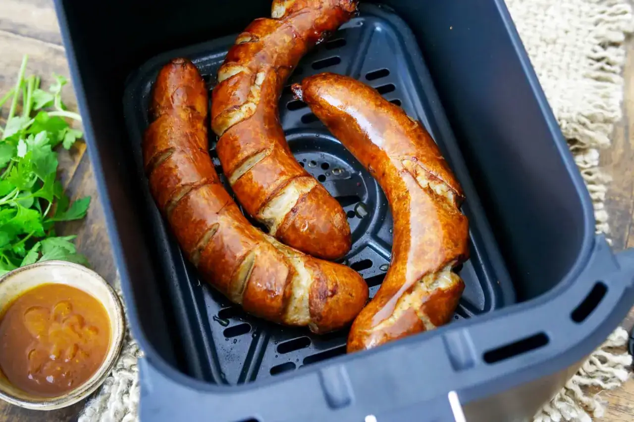 Drei goldbraune Bratwürste im Airfryer, bereit zum Genießen. Daneben eine Schale mit Sauce und frische Petersilie.