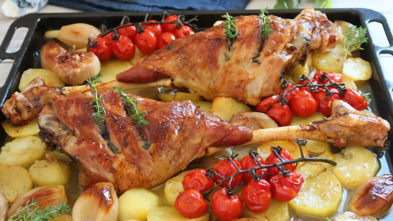 Dos jarretes de cordero asados con patatas, tomates cherry y cebolla, decorados con romero fresco.