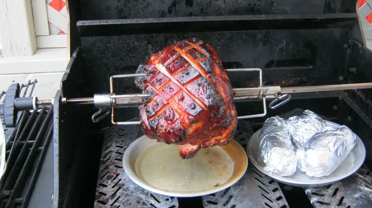 Jambon à la broche pour 50 personnes, doré et grillé, avec des pommes de terre en papillote sur le côté.