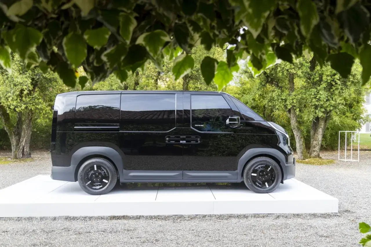 Un elegante minivan elettrico nero, con finestrini oscurati, esposto su una piattaforma bianca in un giardino lussureggiante.