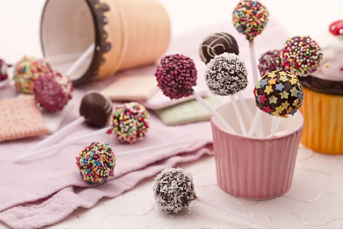Bunte cake pops biskuit, verziert mit Streuseln, Kokosraspeln und Sternen, liegen auf einer rosa Decke.