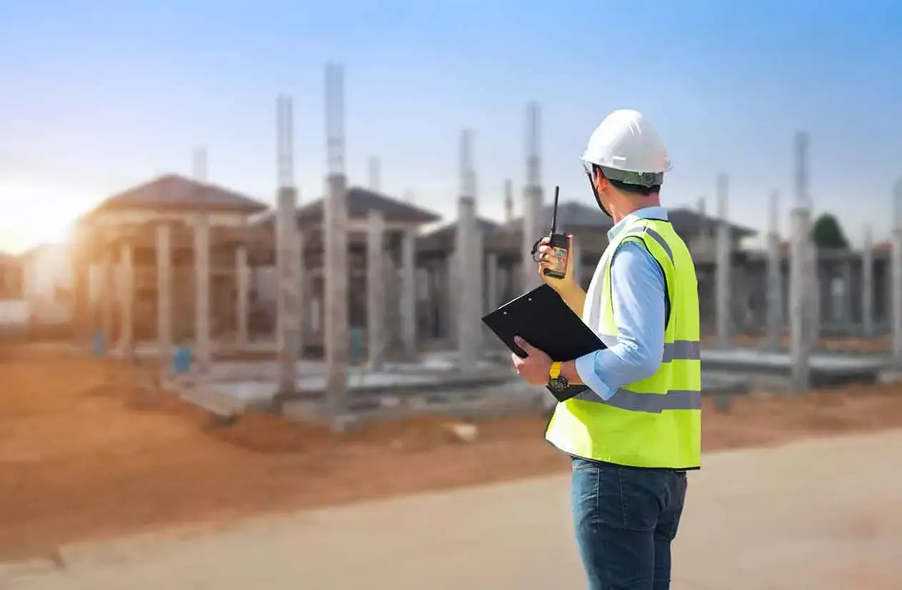 Un capomastro con walkie-talkie e cartella supervisiona un cantiere edile. Il suo ruolo &egrave; fondamentale per il buon esito del progetto.