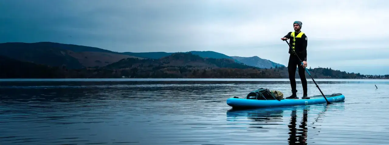 Mężczyzna w kamizelce ratunkowej i czapce stoi na niebieskim SUP-ie, gotowy do sup touring po spokojnym jeziorze. W tle góry i lasy.