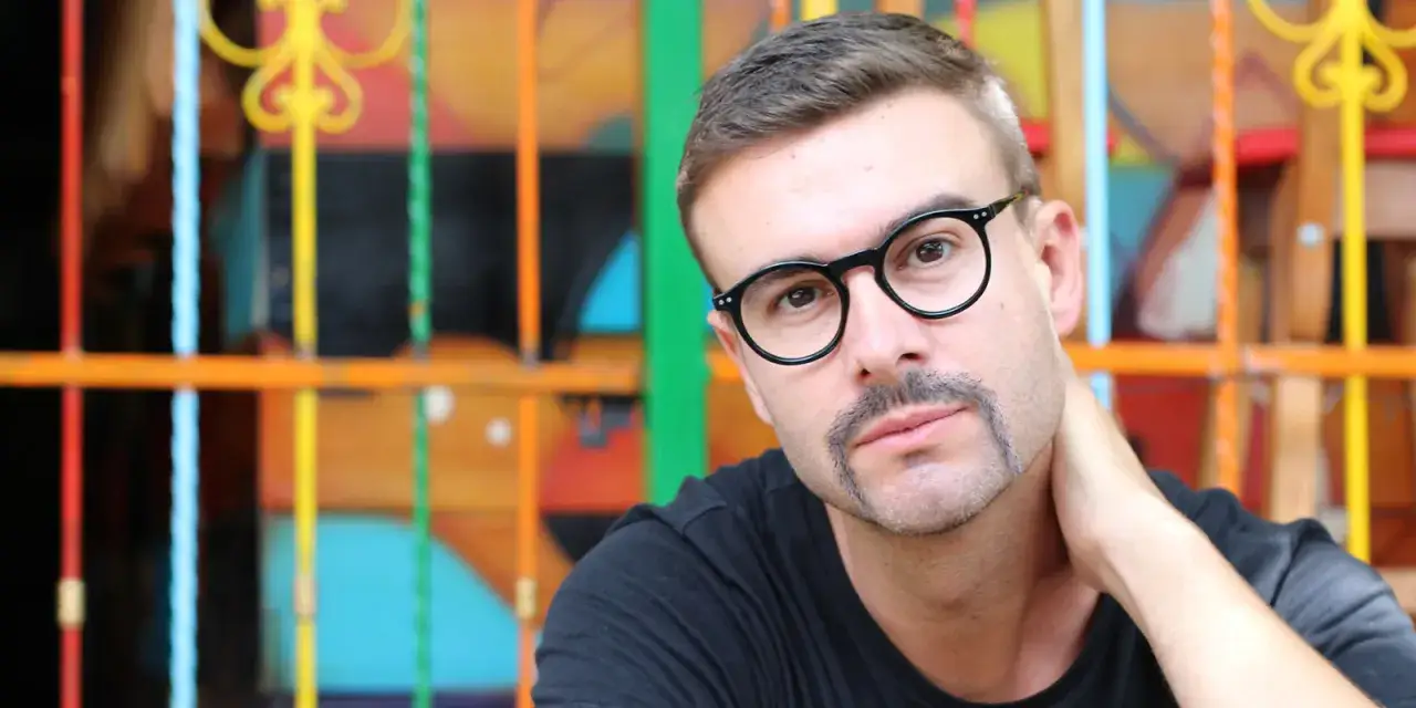 Hombre con gafas y un distintivo bigote herradura, apoyado en una colorida reja.