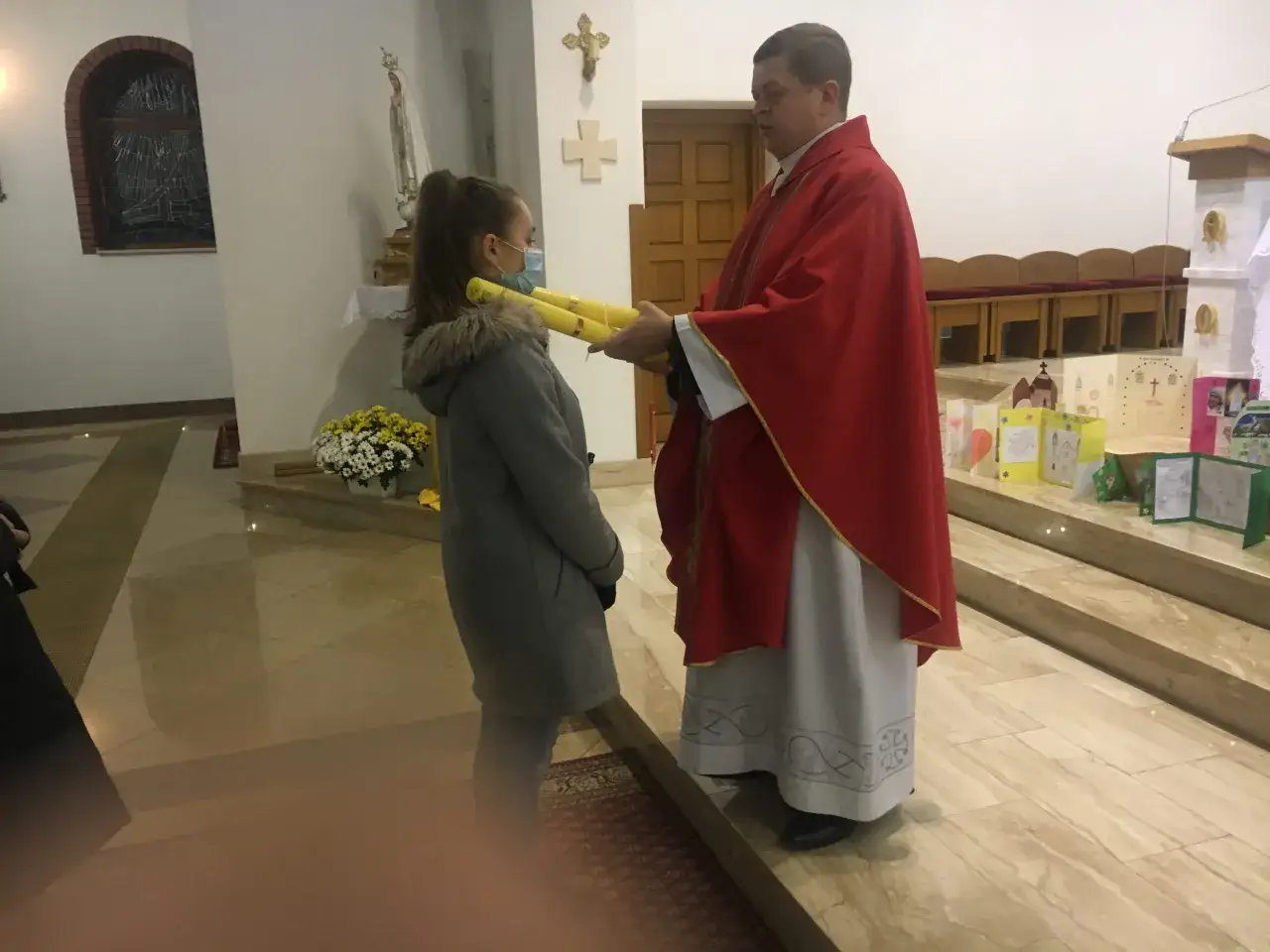 Kapłan w czerwonej dalmatyce wręcza dziewczynce świece. Piękne życzenia dla kapłana od parafian.