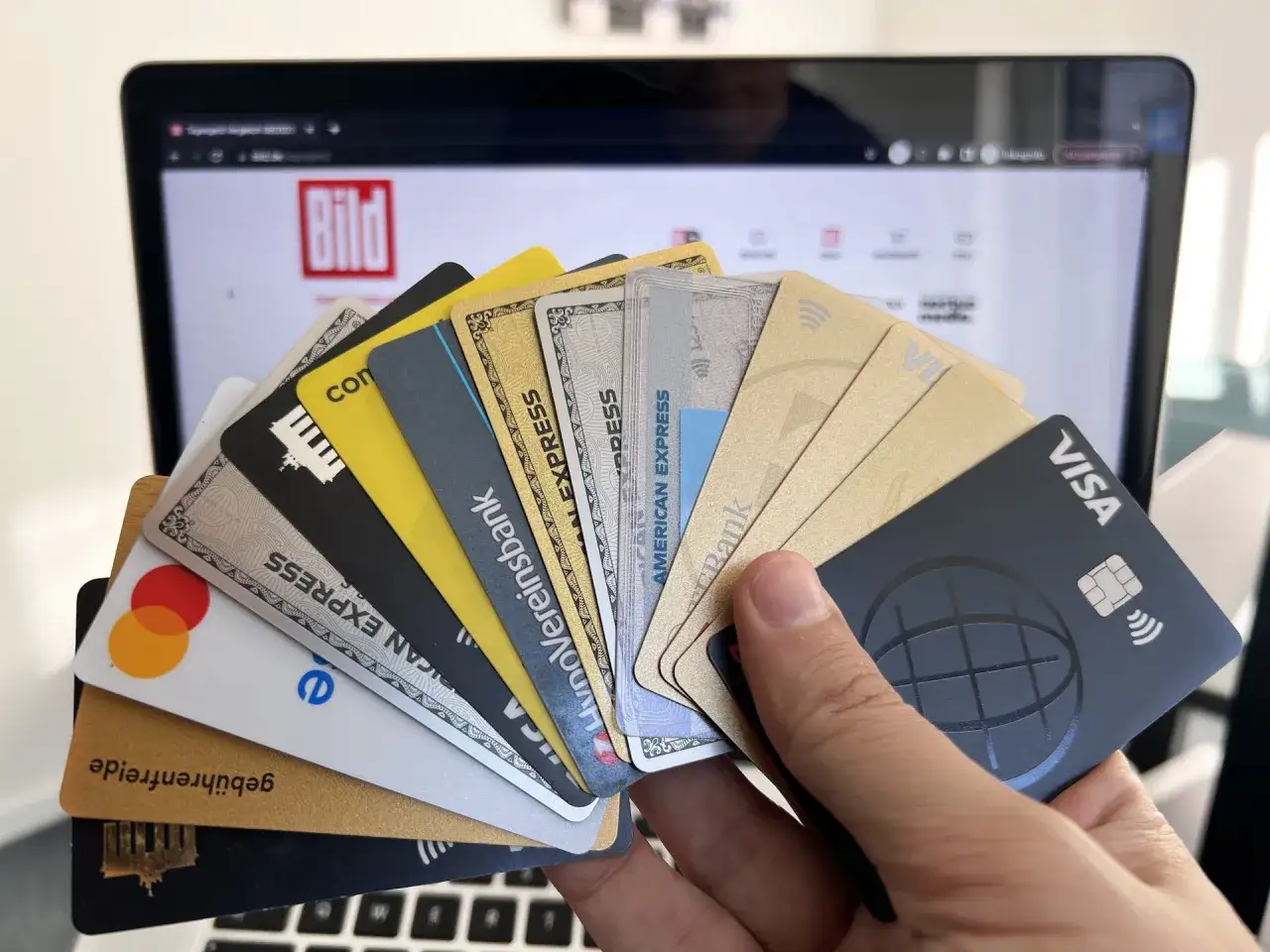 Hand h&auml;lt F&auml;cher von Kreditkarten, darunter Visa und Mastercard, vor einem Laptop mit Bild.de.
