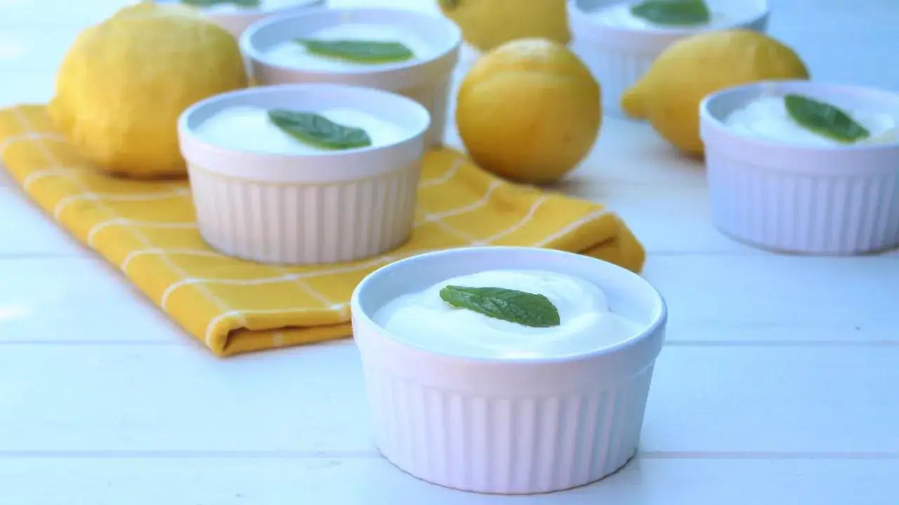 Delicioso mousse de limón mambo en ramequines blancos, adornado con hojas de menta fresca. Limones amarillos y un paño a cuadros completan la escena.