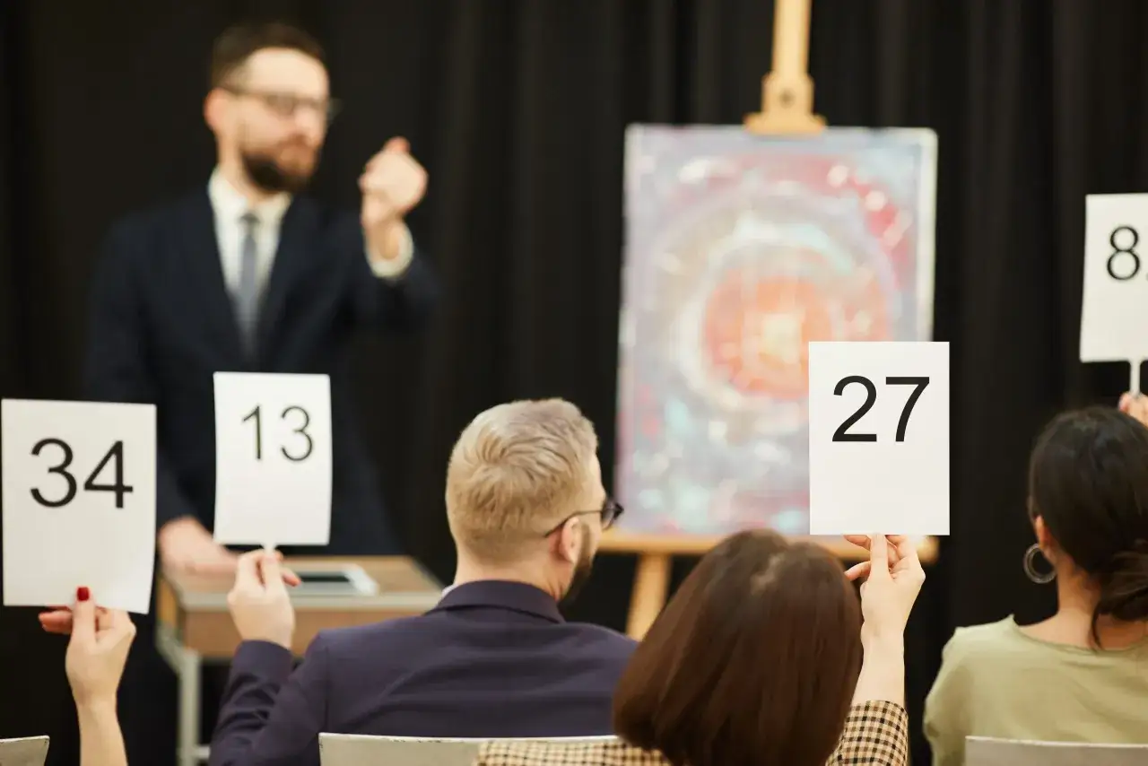 Licytacja dzieła sztuki. Aukcjoner zachęca do podbijania cen, a widzowie podnoszą tabliczki z numerami. Wśr&oacute;d nich jest potencjalny mecenas sztuki.