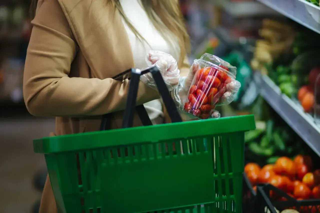 Kobieta w rękawiczkach wkłada pomidorki do zielonego koszyka w holenderskich sklepach.