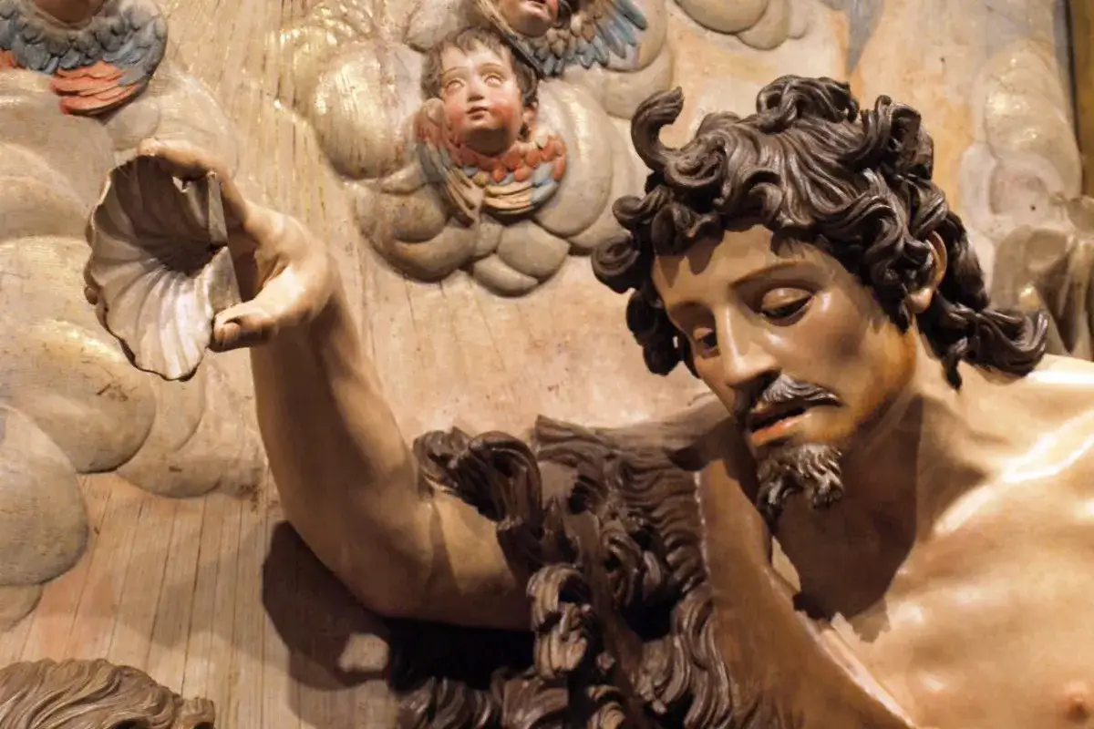 Detalle de escultura barroca: figura masculina con barba y cabello rizado sostiene una concha. Ángeles infantiles observan.