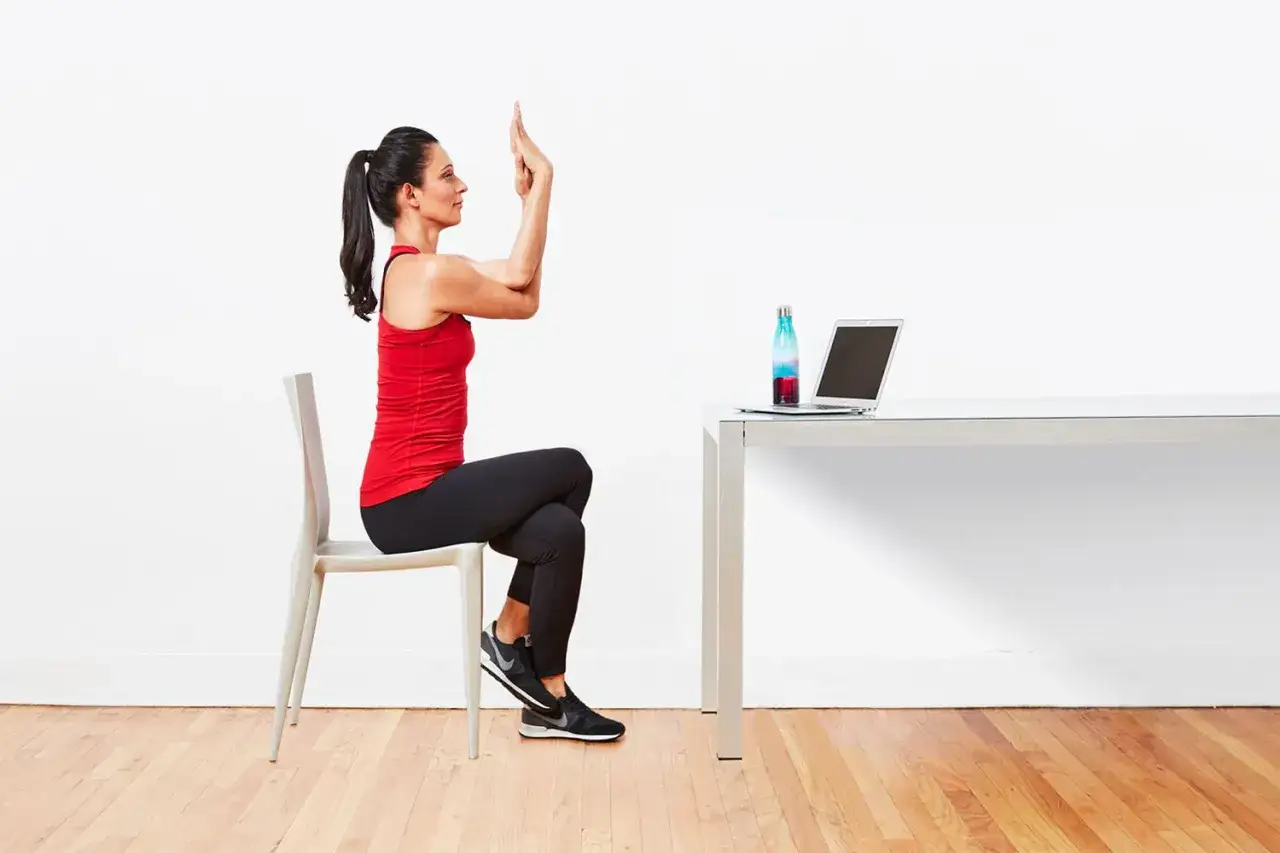 Donna esegue yoga da seduti, con le braccia incrociate in posizione di Garuda. Sul tavolo, un laptop e una borraccia.