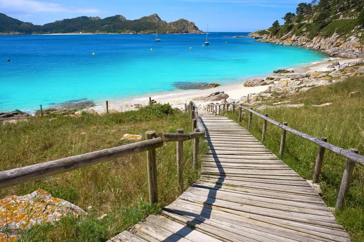Camino de madera hacia una playa paradisíaca, con aguas turquesas y montañas al fondo. Un ejemplo de las 20 mejores playas de Galicia.