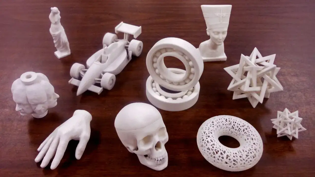 Różnorodne modele wydrukowane na drukarce 3d: czaszka, dłoń, bolid F1, łożyska, popiersie Nefertiti, wielogłowa postać, figurka i geometryczne bryły.