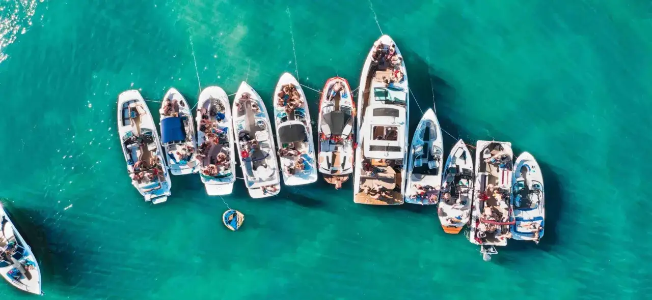 Grupa łodzi cumuje blisko siebie na turkusowej wodzie, tworząc imprezowy prom majorka ibiza. Ludzie relaksują się na pokładach, ciesząc się słońcem i morzem.