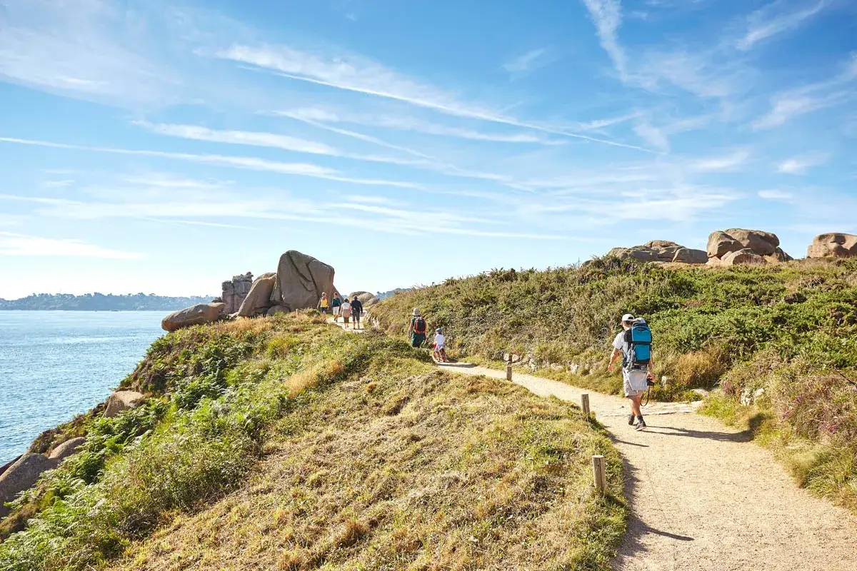 Des randonneurs profitent du soleil pour se balader sur un sentier côtier bordé de rochers et de végétation luxuriante.