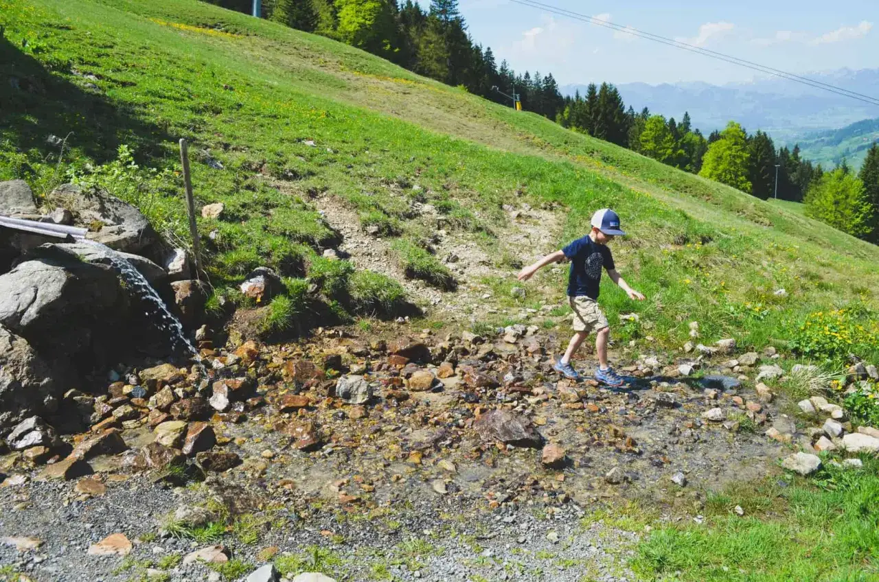 Ein Junge balanciert über Steine in einem Bachbett. Ein tolles Erlebnis beim **Bad Hindelang wandern mit Kindern** im Grünen.