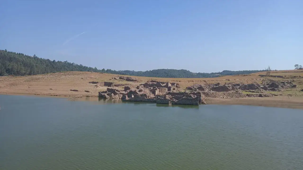 Ruinas de un pueblo sumergido emergen del embalse de Portodemouros, con colinas boscosas al fondo bajo un cielo azul.