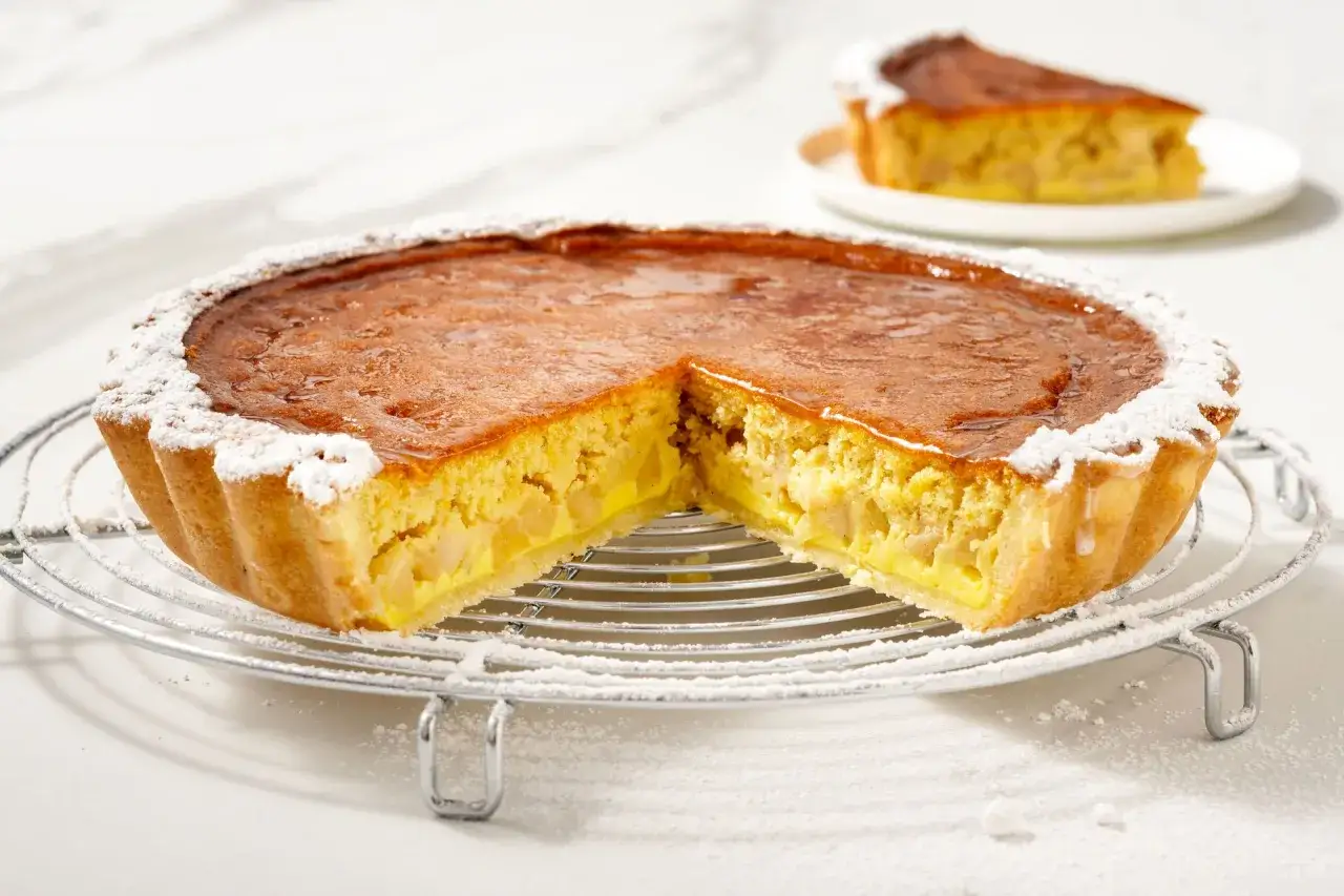 Una deliziosa torta di mele con crema pasticcera, cosparsa di zucchero a velo, su una griglia metallica. Una fetta &egrave; gi&agrave; servita su un piattino.