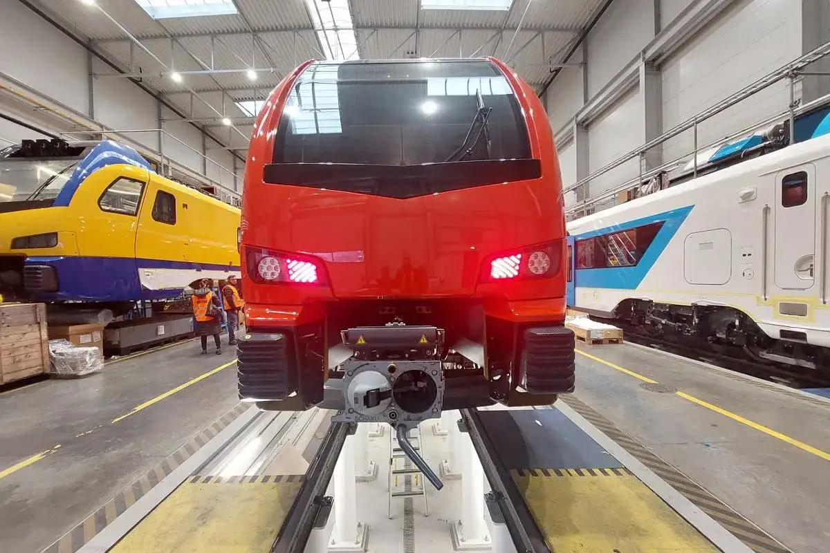 Czerwony pociąg Stadler Rail w warsztacie, obok żółto-niebieskiego i białego składu.