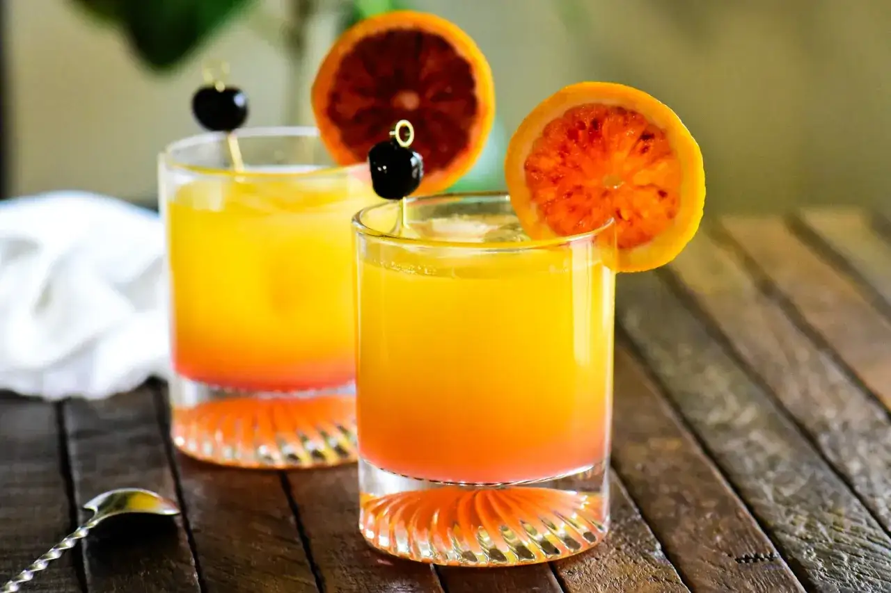 Dwa drinki tequila sunrise z pomarańczą i wiśnią na patyczku, na drewnianym stole.
